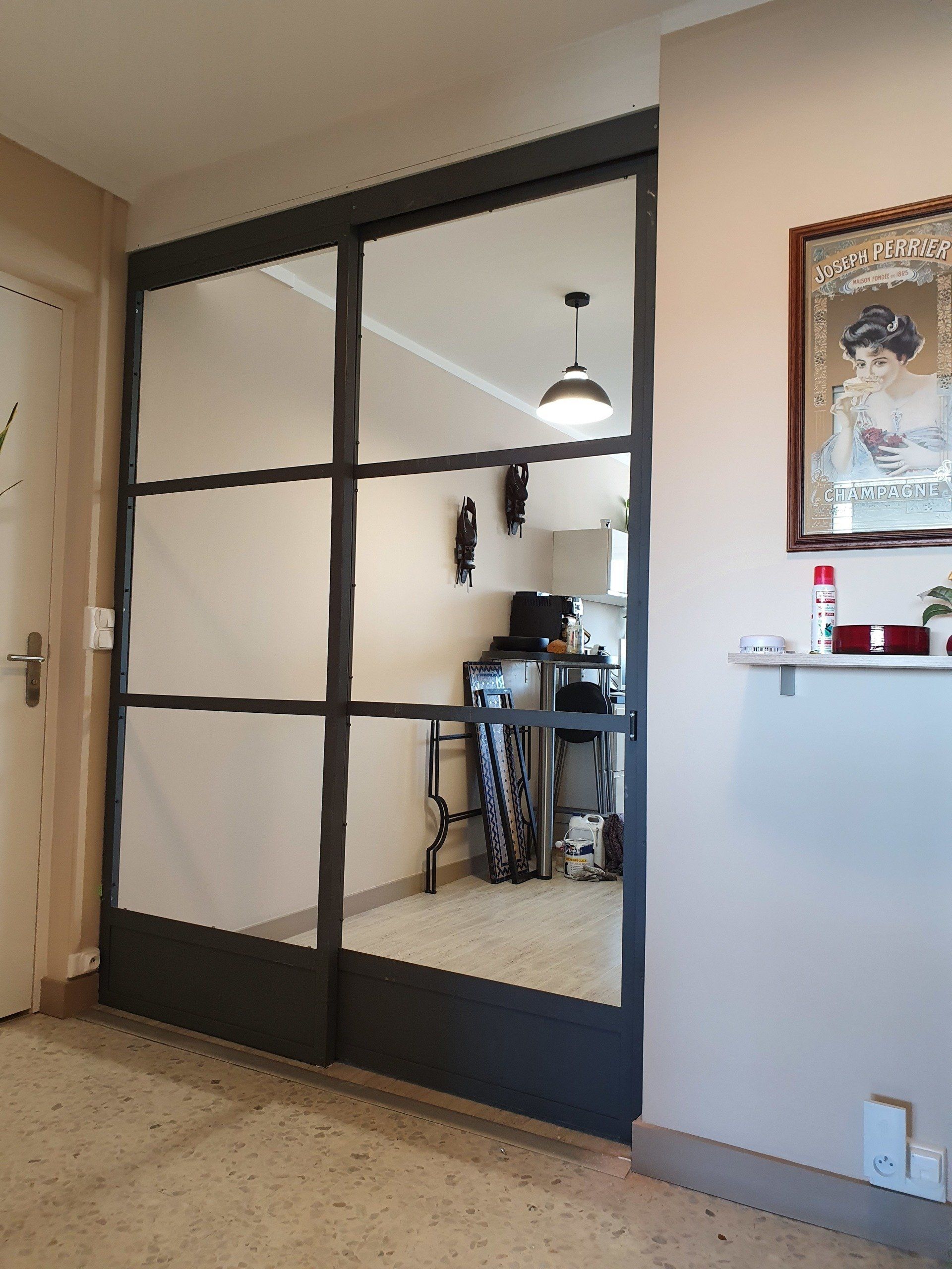 Des portes coulissantes en verre à cadre métallique noir reflètent une pièce avec un bureau et une décoration.