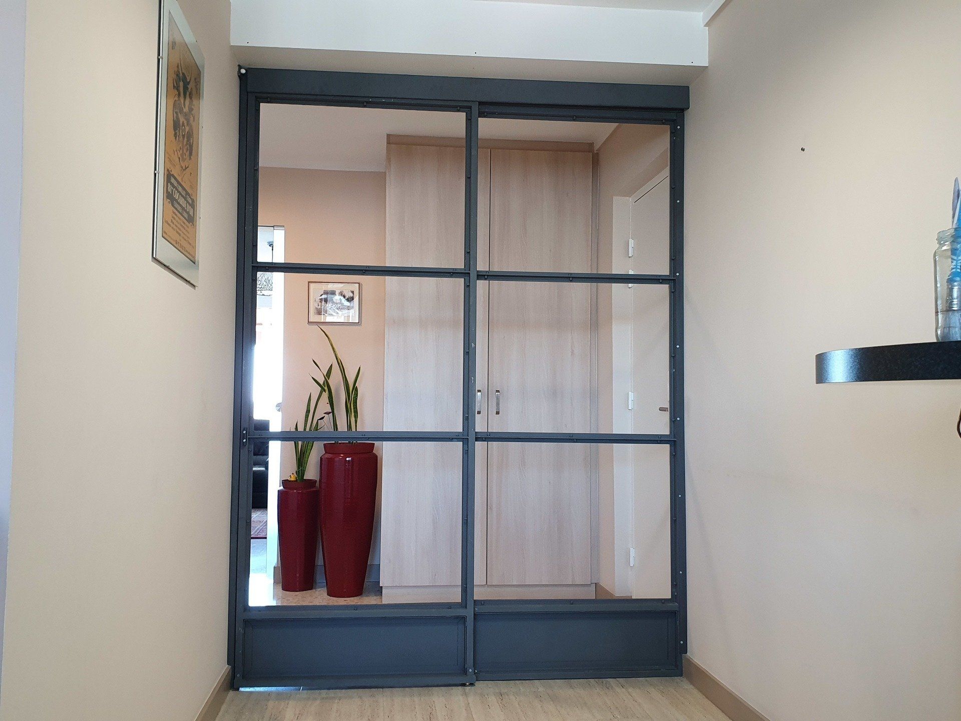 Porte vitrée aux encadrements gris foncé. Derrière la vitre, deux grands vases rouges contenant des plantes. Murs couleur crème.