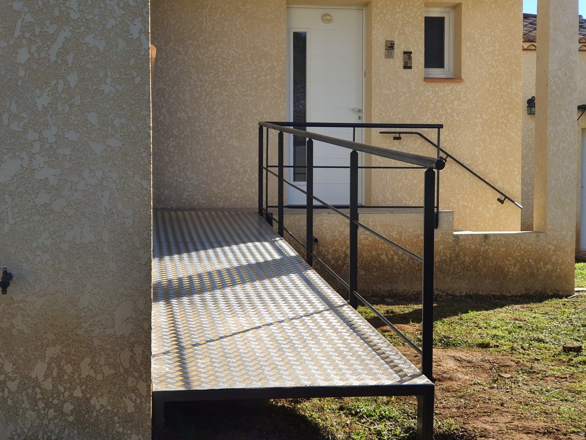 Rampe métallique avec garde-corps menant à une porte. Bâtiment beige, cadre extérieur ensoleillé.