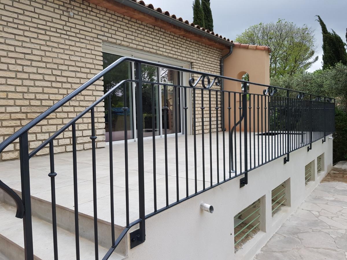 Une balustrade en fer noir entoure un patio avec un escalier descendant. Un bâtiment en briques beiges se dresse à l'arrière-plan.