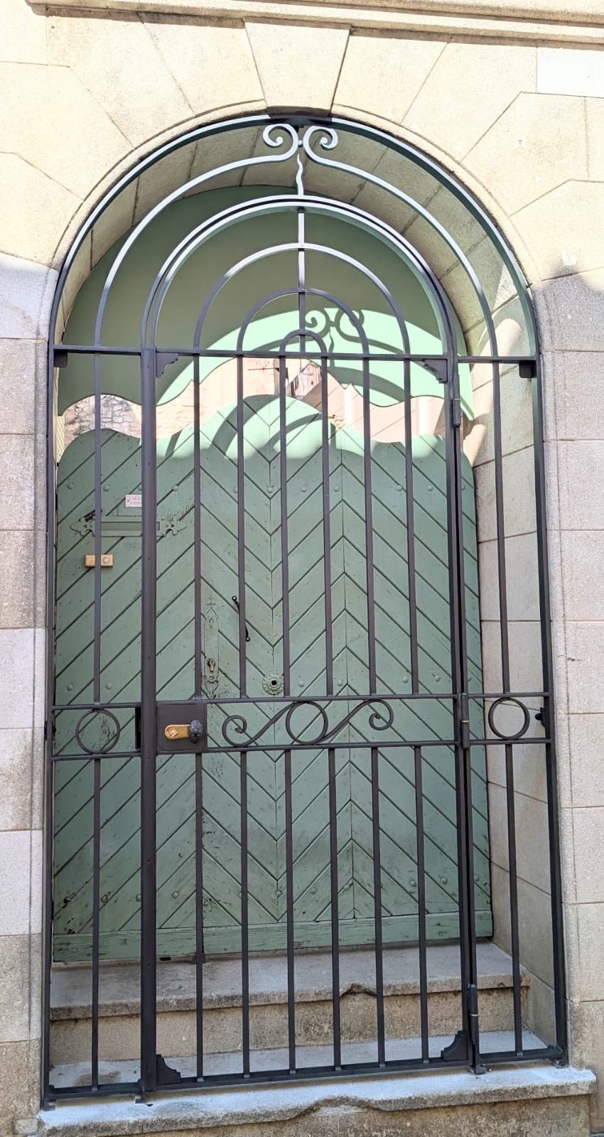 Porte verte derrière un portail en fer forgé noir, encadrée par une arche en pierre.