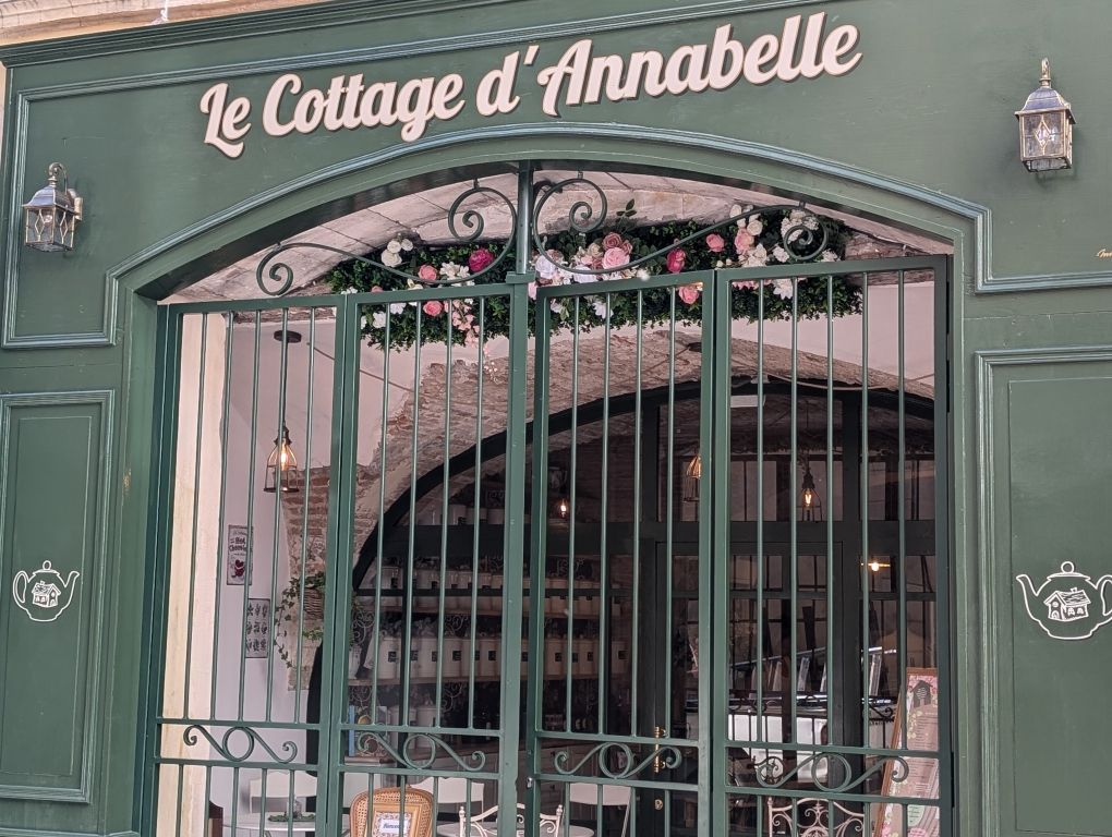 Façade verte avec grille en fer forgé, enseigne « Le Cottage d'Annabelle » au-dessus, arche fleurie et dessins de théières.