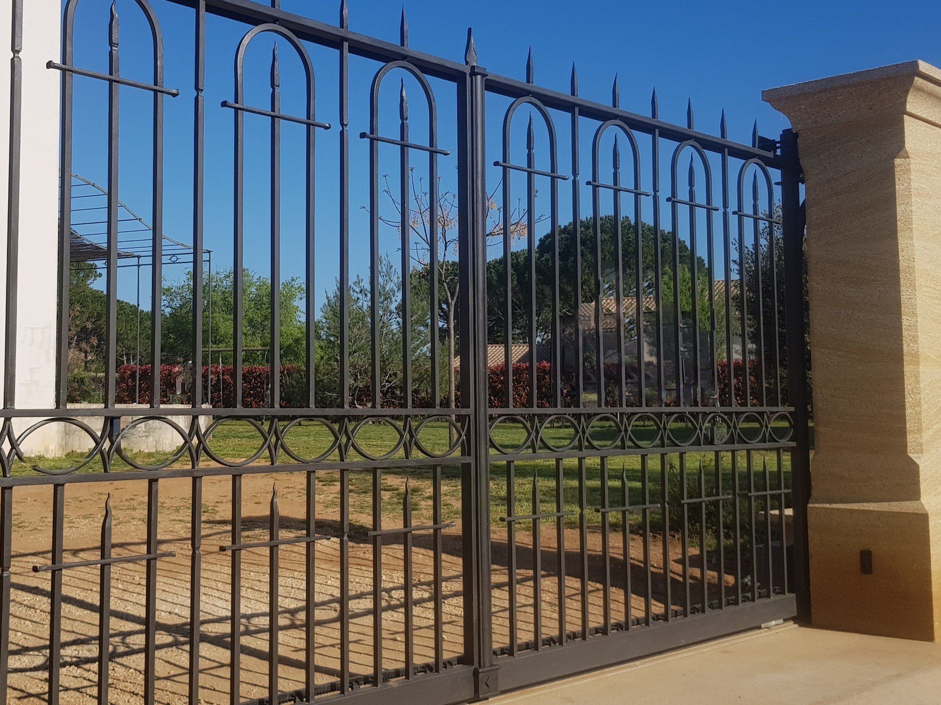 Un portail en fer noir se dresse devant un pilier en pierre beige, offrant une vue sur les arbres et l'herbe au-delà.