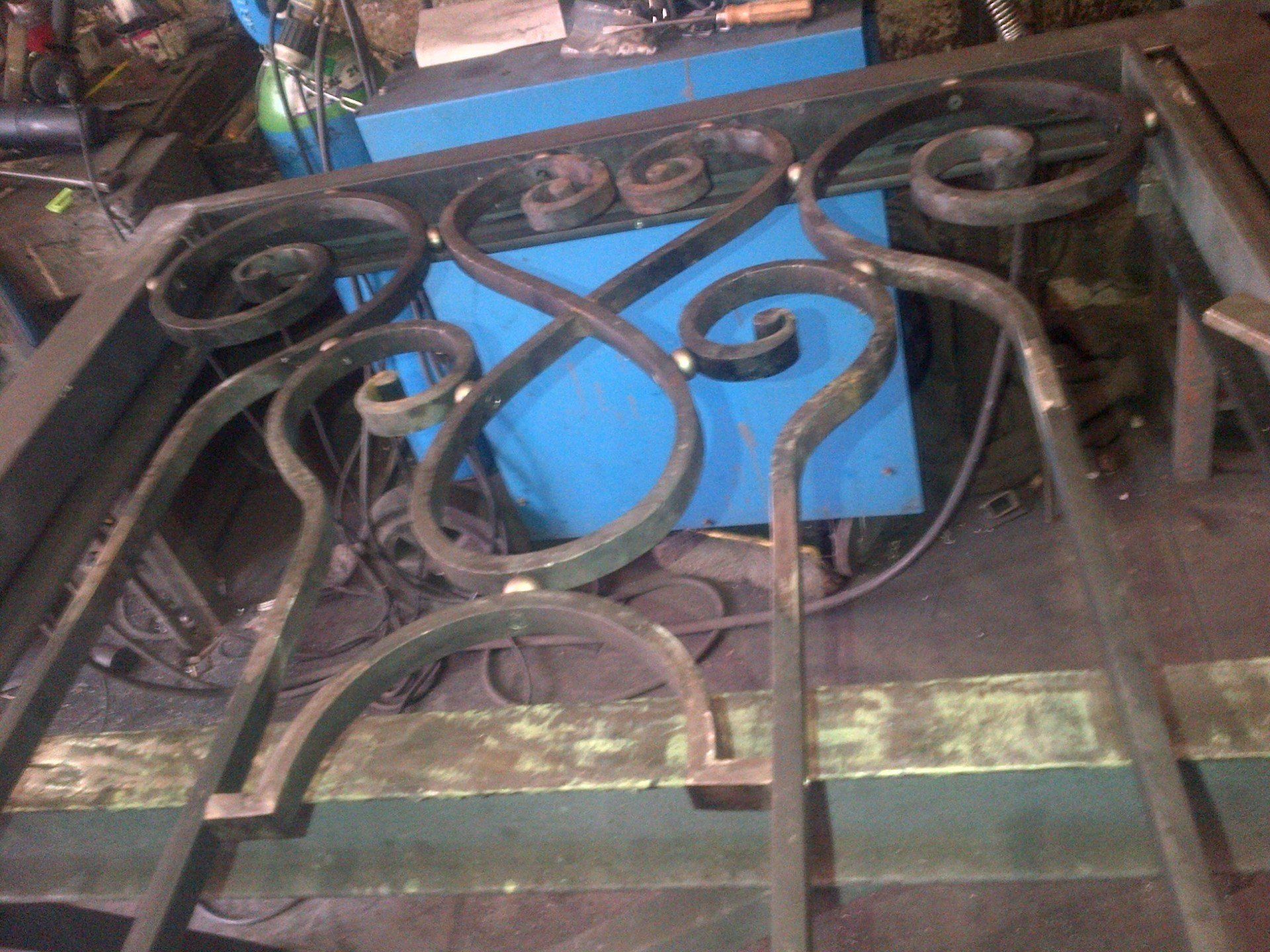 Portail métallique en construction, orné de volutes décoratives, de soudures et d'une machine bleue en arrière-plan.