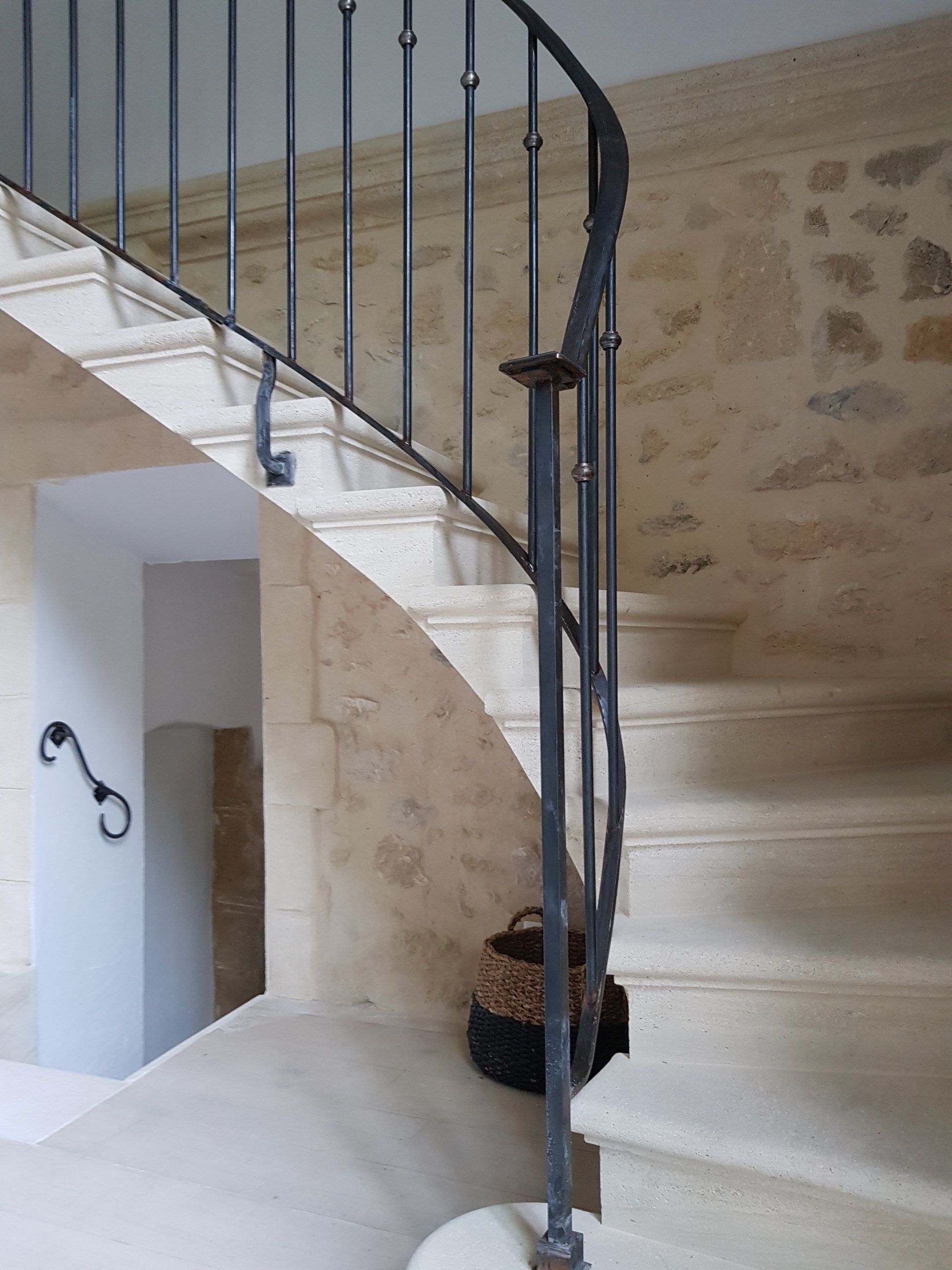 Escalier en pierre avec rampe en fer forgé adossé à un mur de pierre. Un panier tressé est posé sur les marches.