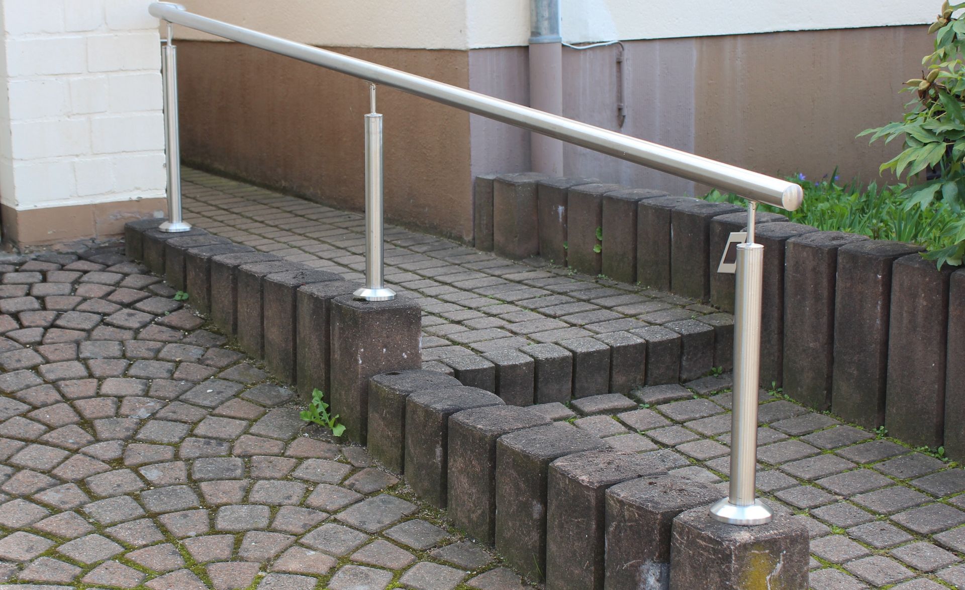 Ein Edelstahlgeländer auf einem Kopfsteinpflasterweg neben einem Gebäude.