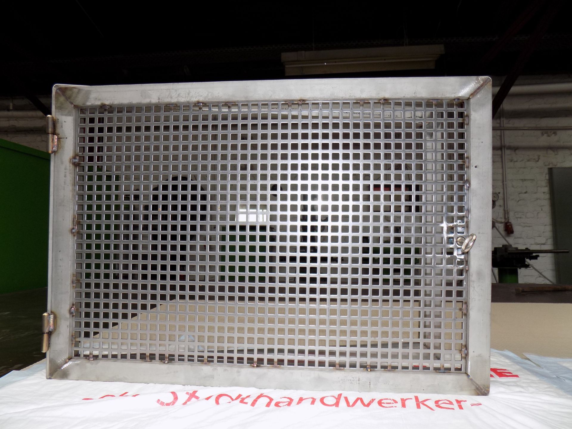 Ein Edelstahlgitter liegt auf einem Stück Papier mit der Aufschrift jx-handwerker