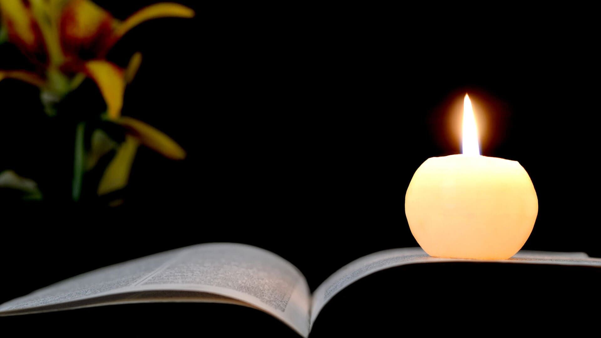 Livro aberto com uma vela acesa em cima, flores à esquerda, tudo sobre um fundo preto.