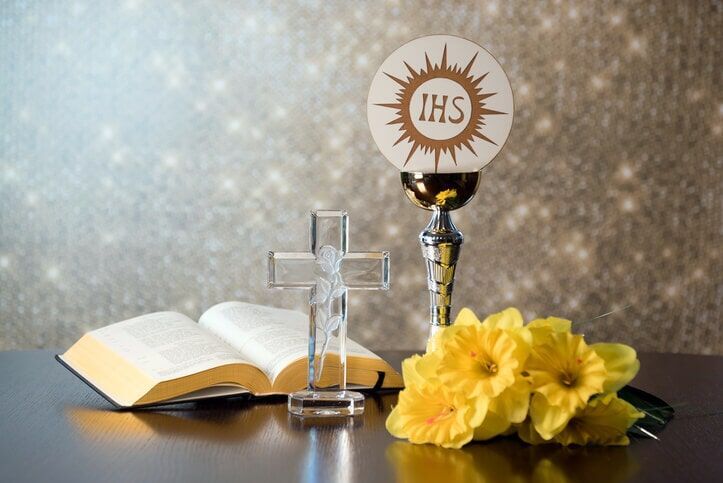 Cálice com hóstia, Bíblia aberta, cruz e flores amarelas sobre uma mesa, com fundo brilhante.