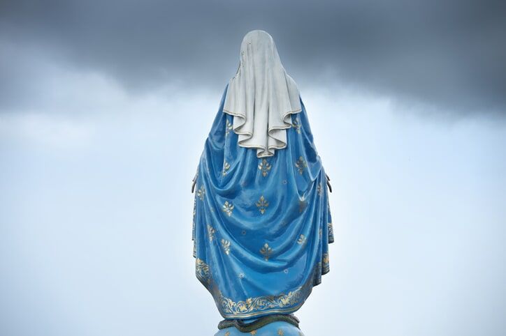 Vista posterior de uma estátua de Maria vestindo um manto azul e um véu branco contra um céu nublado.