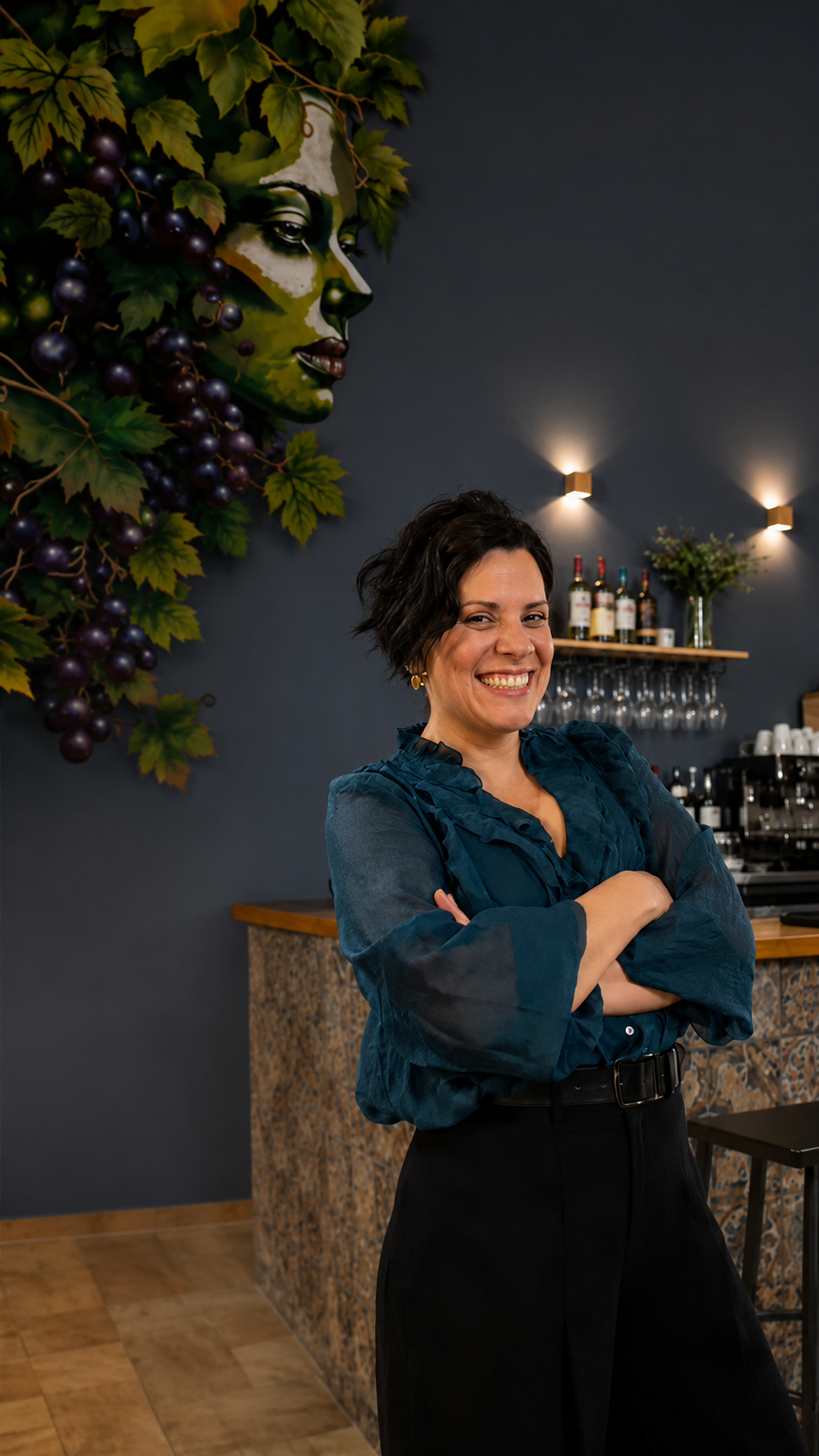 Una mujer con blusa color turquesa aparece de pie, sonriendo y con los brazos cruzados, en un bar con poca luz