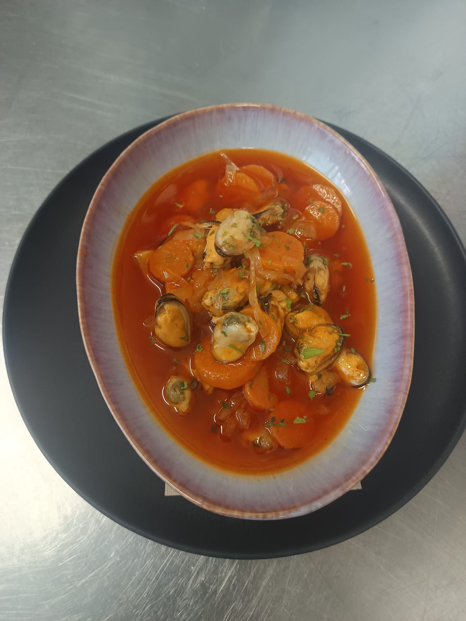 Sopa roja con almejas y hierbas en un tazón ovalado blanco sobre un plato oscuro.