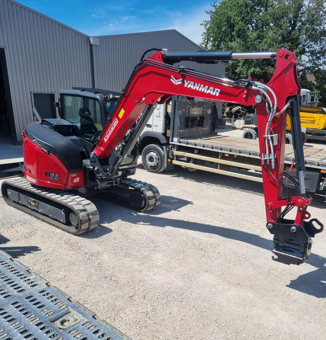 Minipelle Yanmar rouge sur gravier, garée devant un bâtiment et un camion, en extérieur.