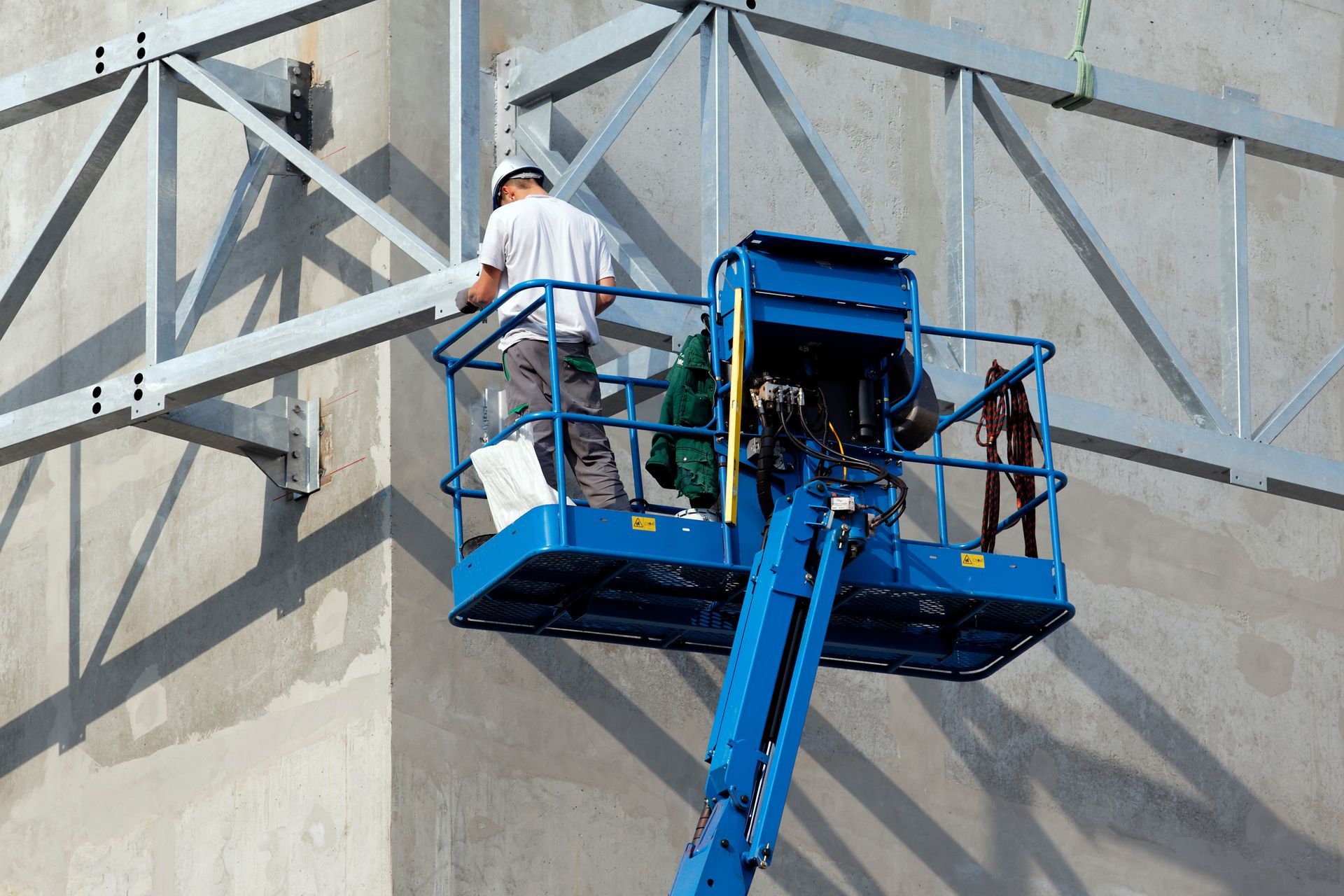 Un ouvrier du bâtiment, sur une plateforme élévatrice bleue, fixe des poutres métalliques à un mur en béton.
