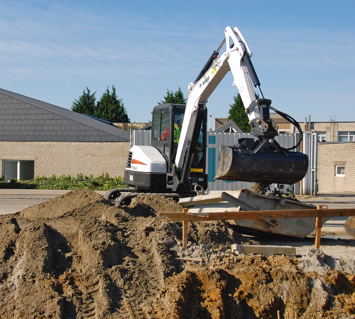 Une petite pelleteuse blanche creuse dans un tas de terre, devant un bâtiment.