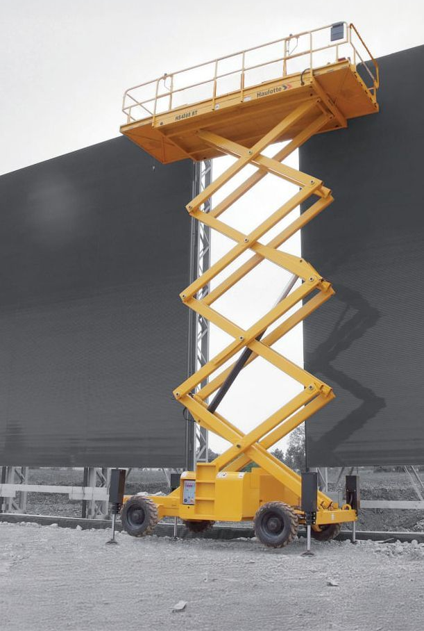 Plateforme élévatrice à ciseaux jaune levée, devant un fond gris, sur un chantier de construction.