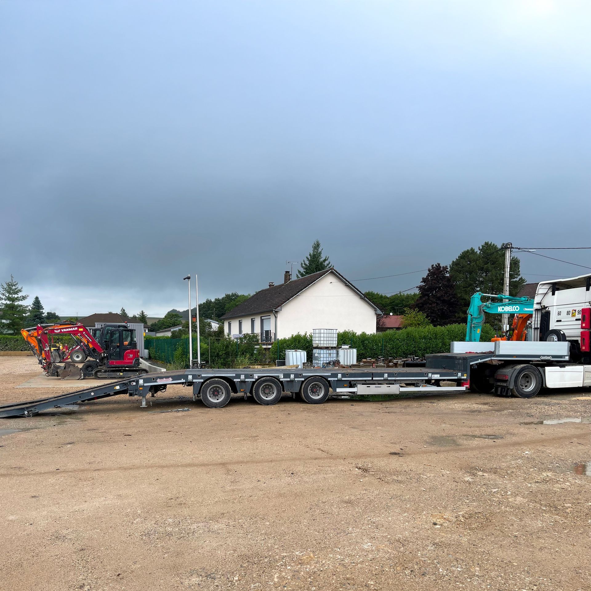 Camion plateau transportant du matériel de chantier stationné sur un terrain gravillonné près de maisons, sous un ciel nuageux.