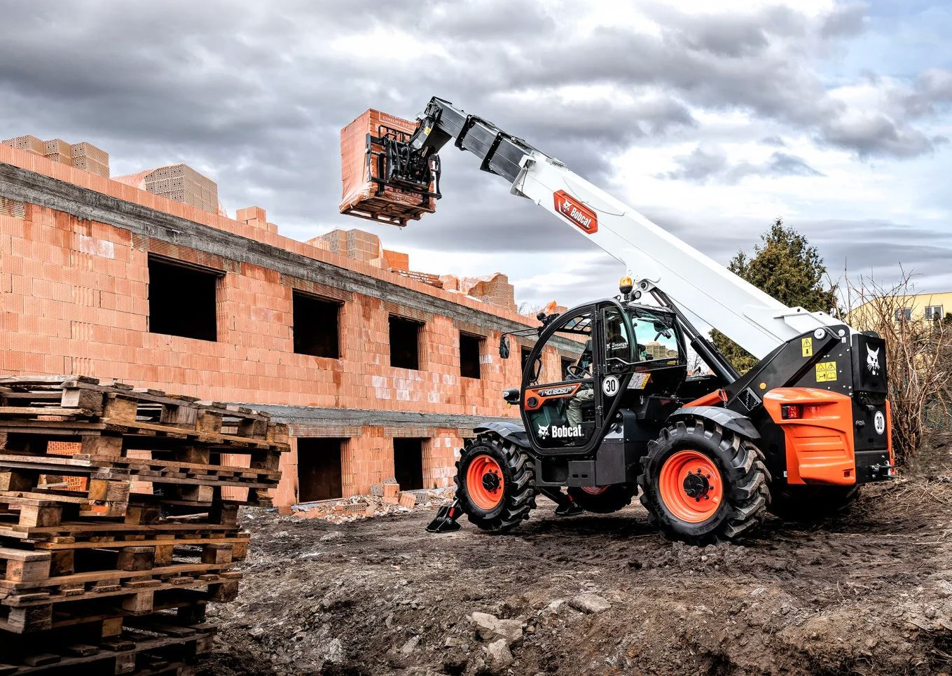 Un chariot télescopique soulève des briques sur un chantier. Engin noir et orange avec flèche blanche déployée.