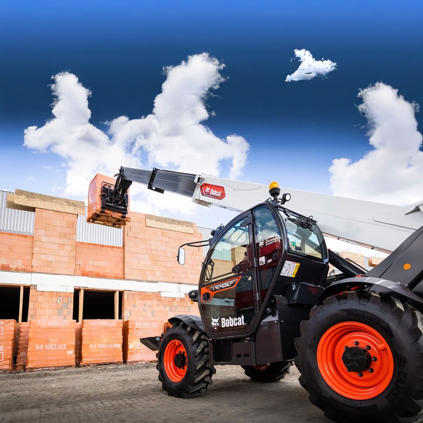 Un chariot télescopique soulève une palette de briques sur un chantier de construction ; ciel bleu et nuages ​​en arrière-plan.
