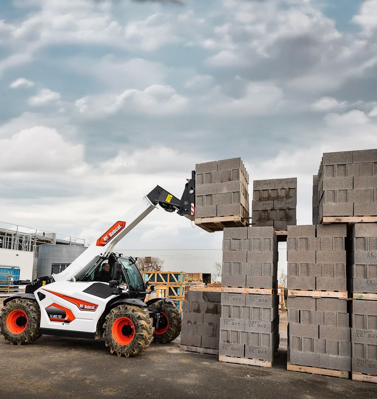 Un chariot télescopique Bobcat soulève des blocs dans une zone industrielle ; machine blanche, pneus orange, ciel nuageux.