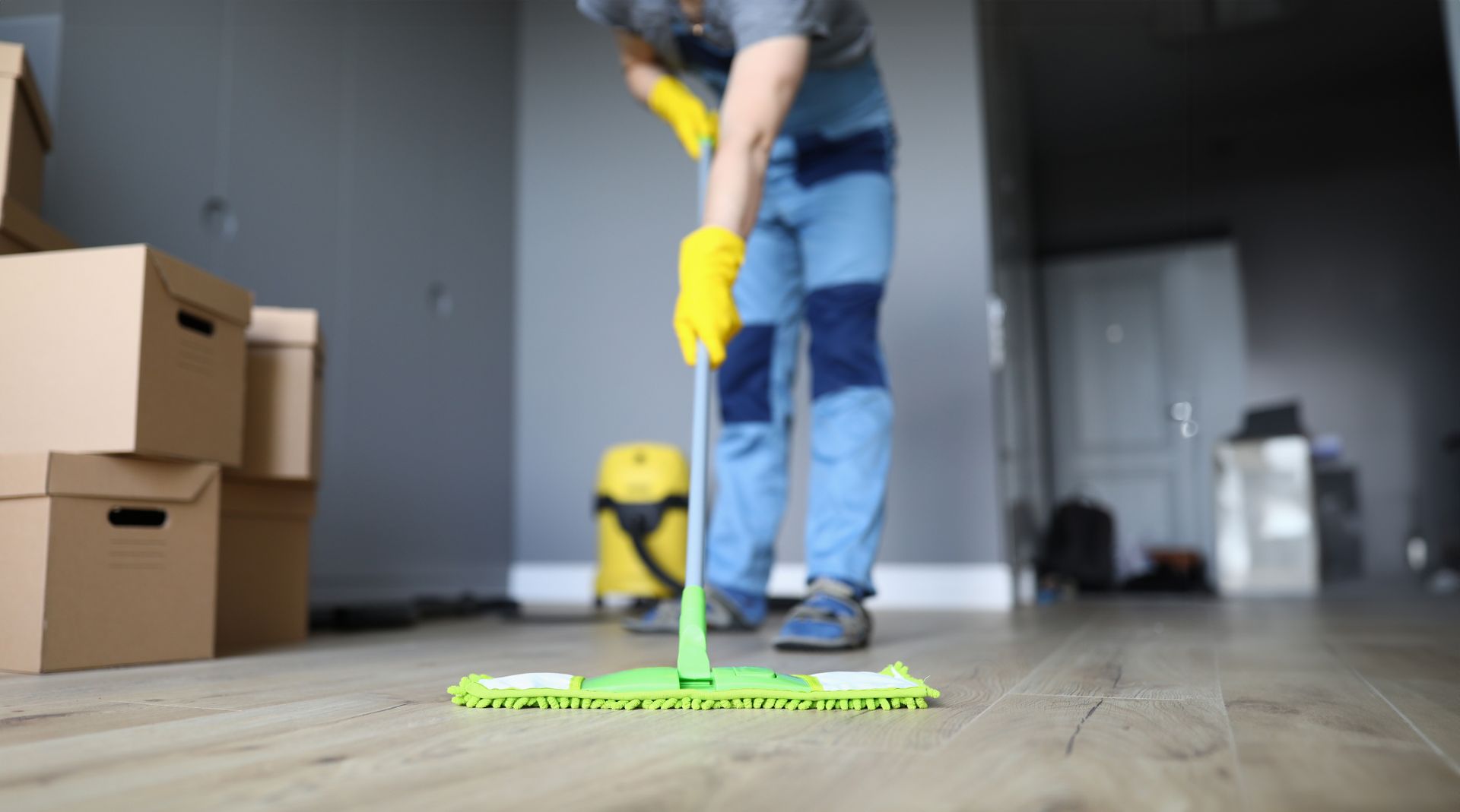 Person in blauer Arbeitskleidung und gelben Handschuhen wischt mit einem hellgrünen Mopp über den Boden in einem Raum.