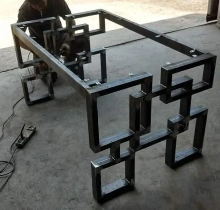 Escultura geométrica de metal negro sobre un suelo de hormigón, con una persona trabajando en ella al fondo.