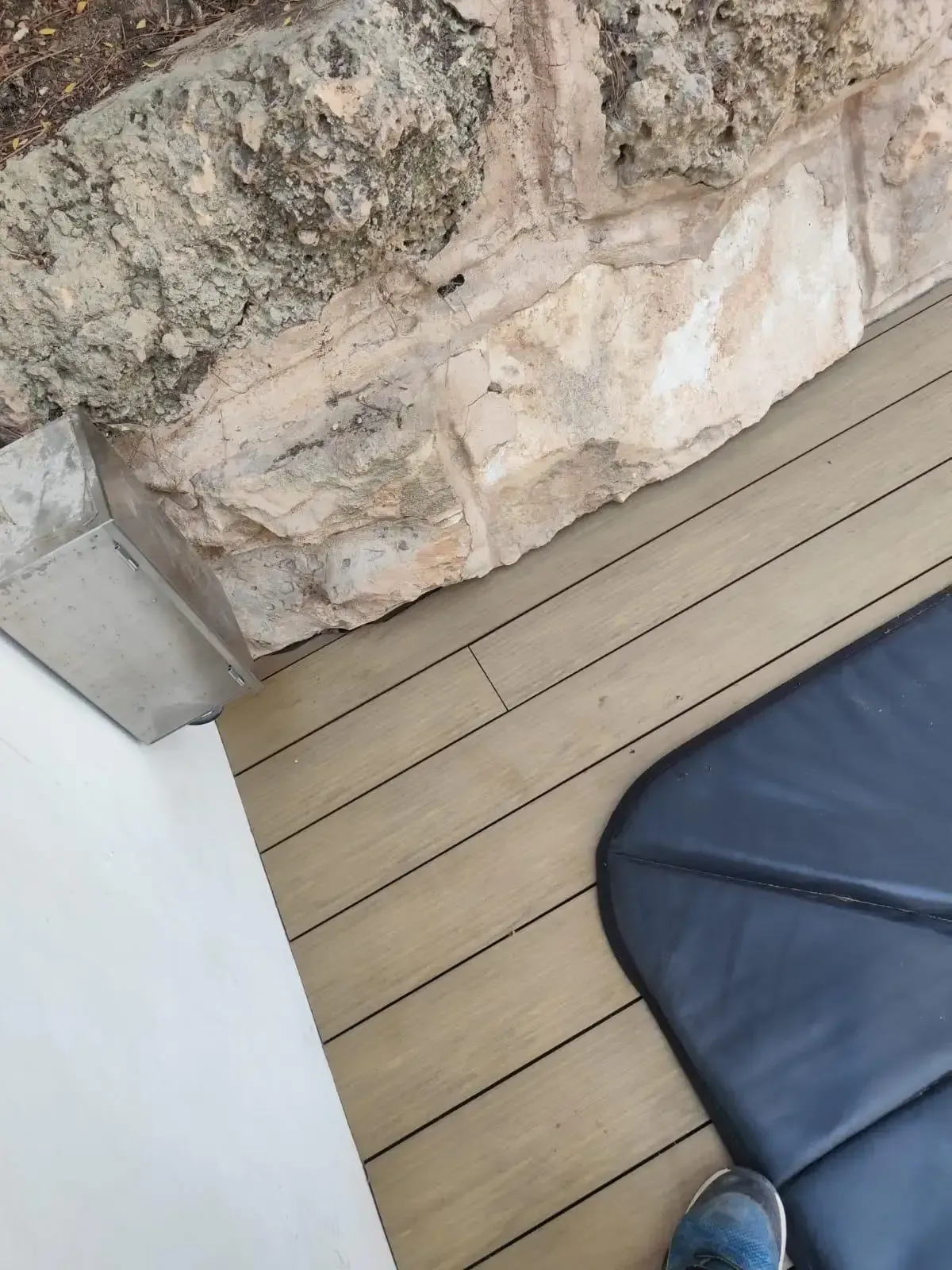 Muro de piedra junto a una tarima de madera clara con un cojín negro en la esquina.