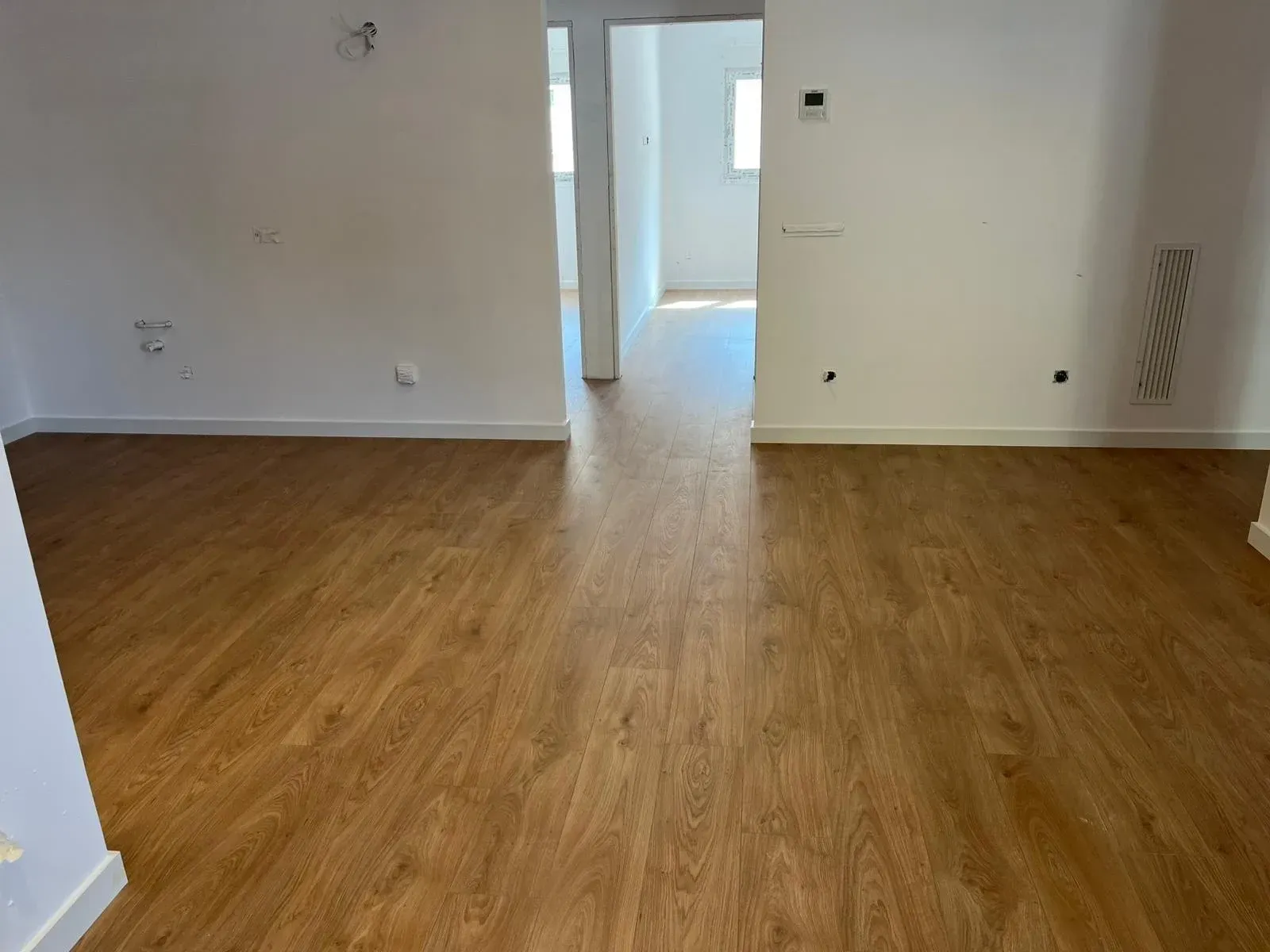 Habitación vacía con suelo de madera y paredes blancas, con vista hacia una puerta y un pasillo.