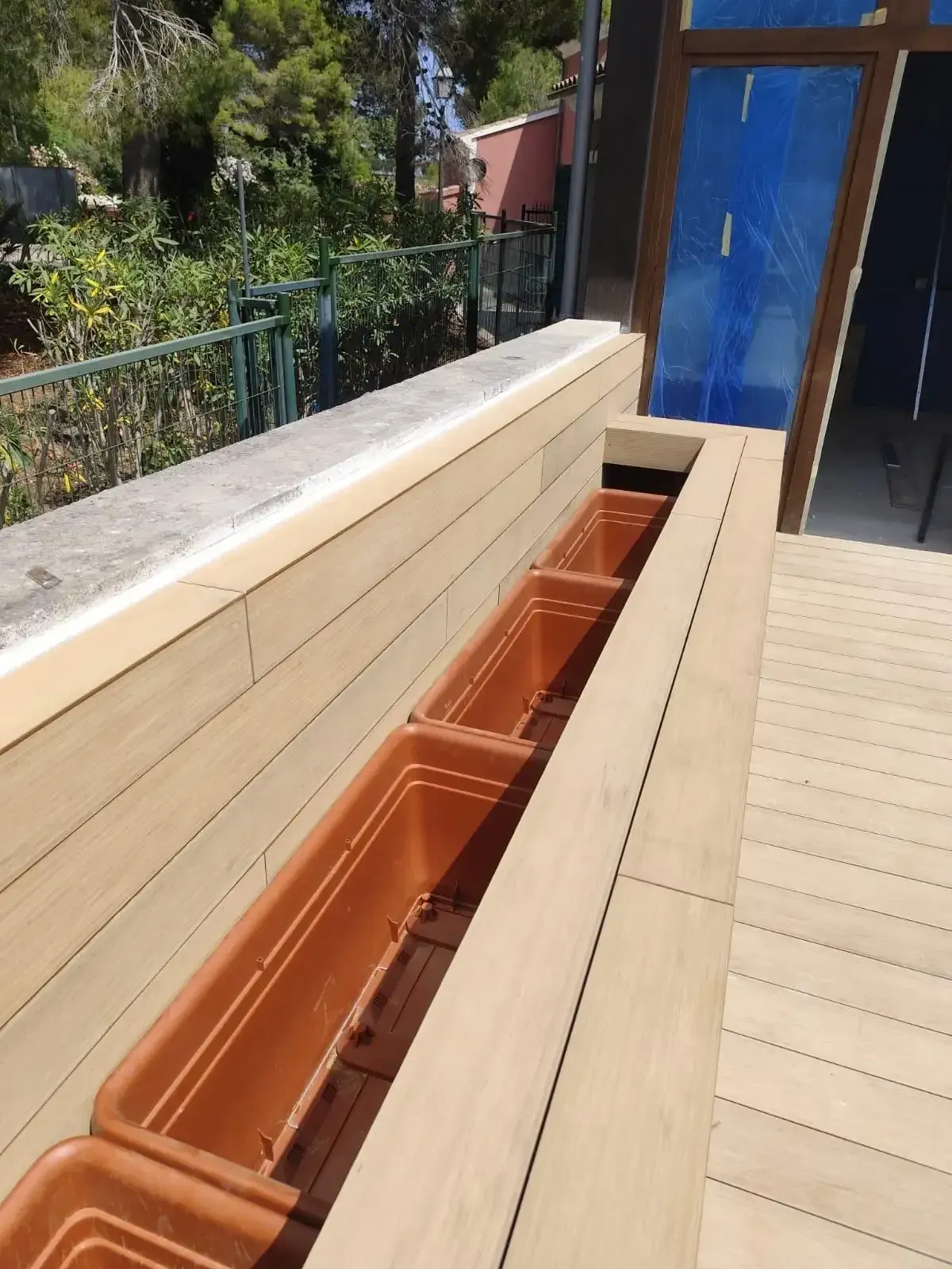 Maceteros largos de madera junto a la barandilla de una terraza, con puertas azules al fondo.