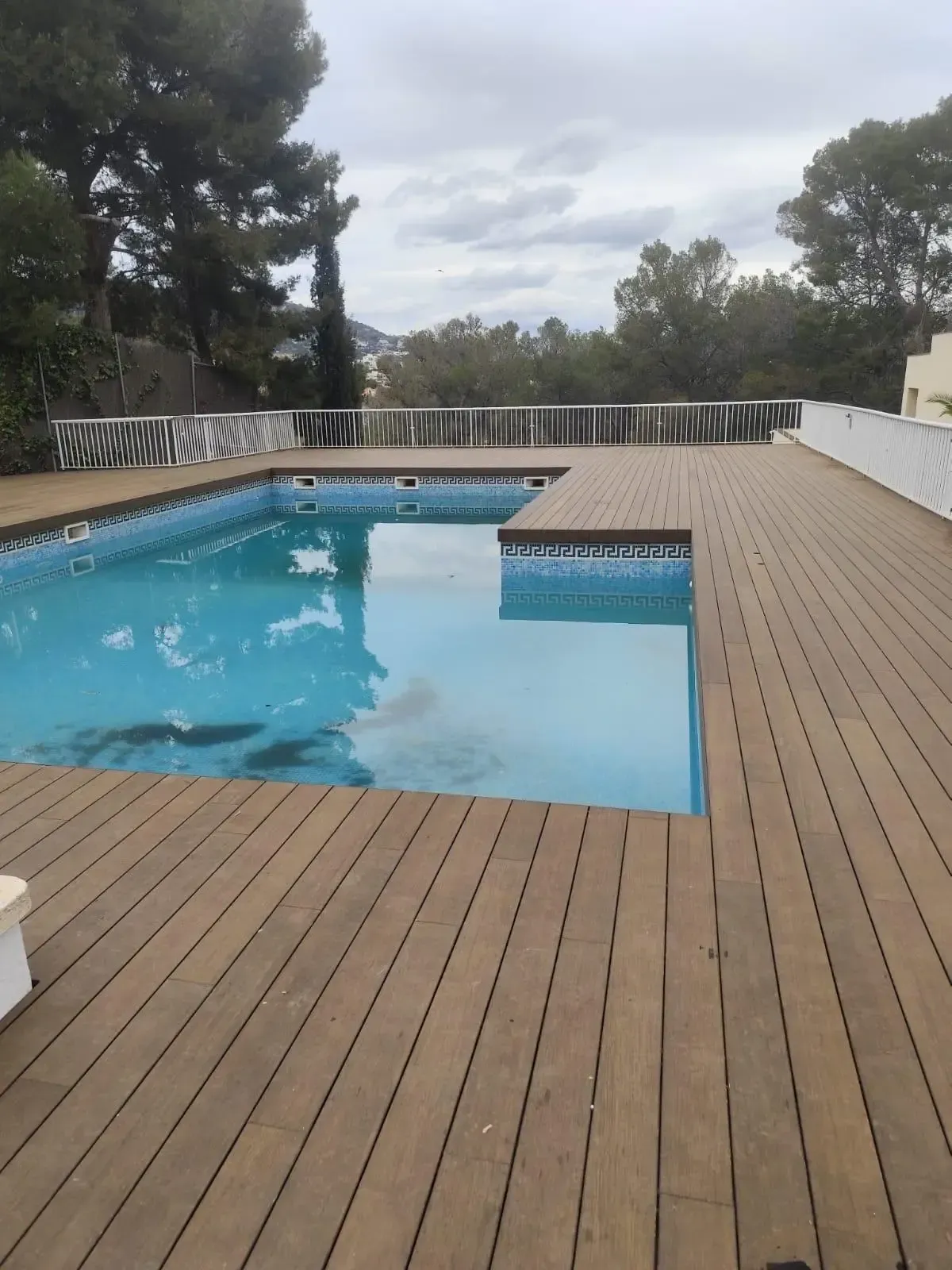 Piscina con terraza de madera y cielo nublado, con vistas a los árboles y al agua.