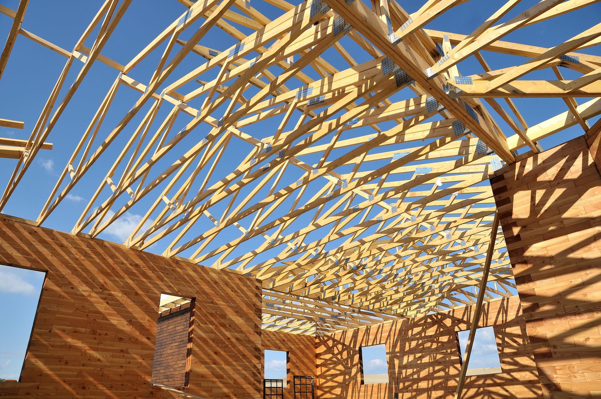 La charpente d'une maison en bois est en construction, on aperçoit un ciel bleu clair à travers le toit.
