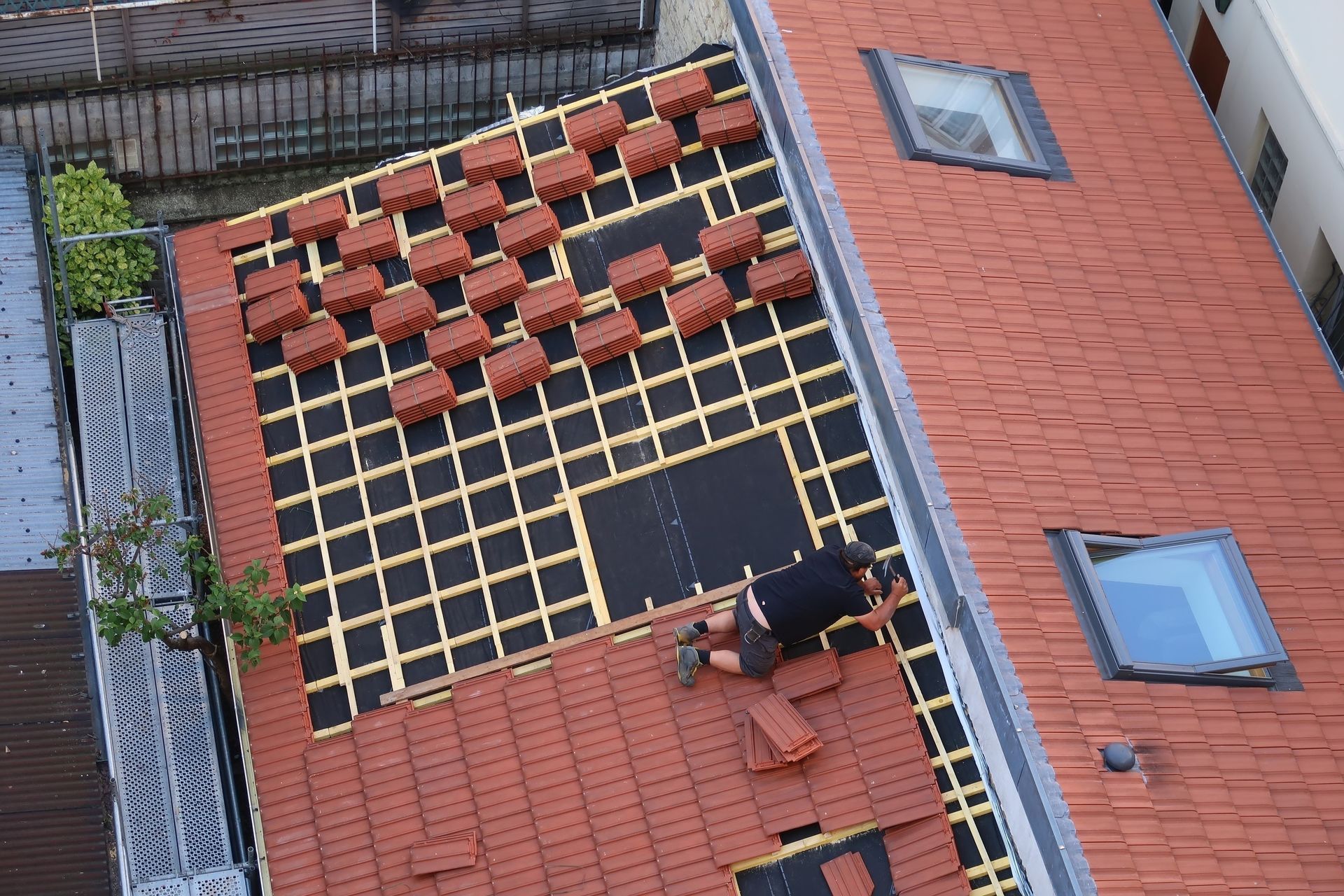 Toit de tuiles rouges baigné de soleil, avec un bâtiment blanc et une cheminée en arrière-plan.