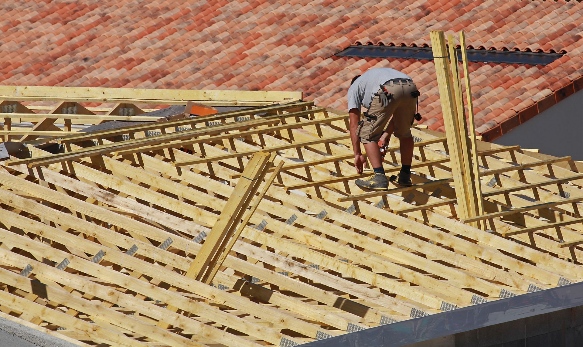 Couvreur sur un toit en construction, utilisant des outils pour installer des poutres en bois. Toiture en tuiles rouges en arrière-plan.