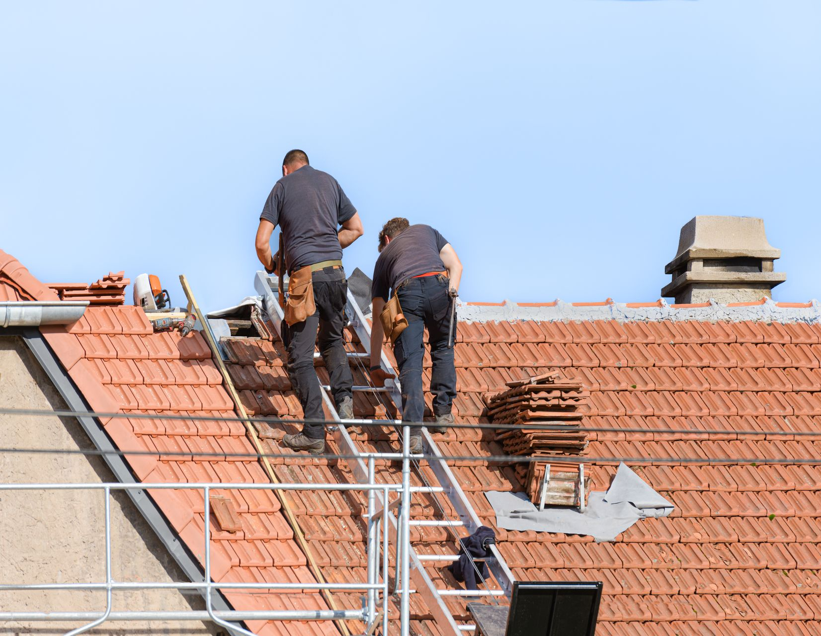 Deux couvreurs sur un toit de tuiles rouges, travaillant avec leurs outils sous un ciel bleu.