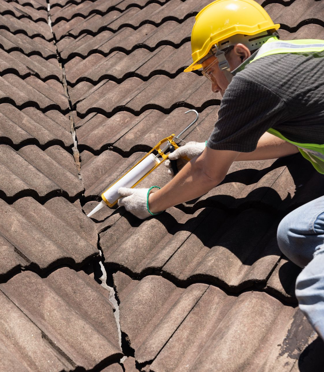 Une personne portant un casque et un gilet de sécurité colmate une fissure dans un toit de tuiles à l'aide d'un pistolet à calfeutrer.