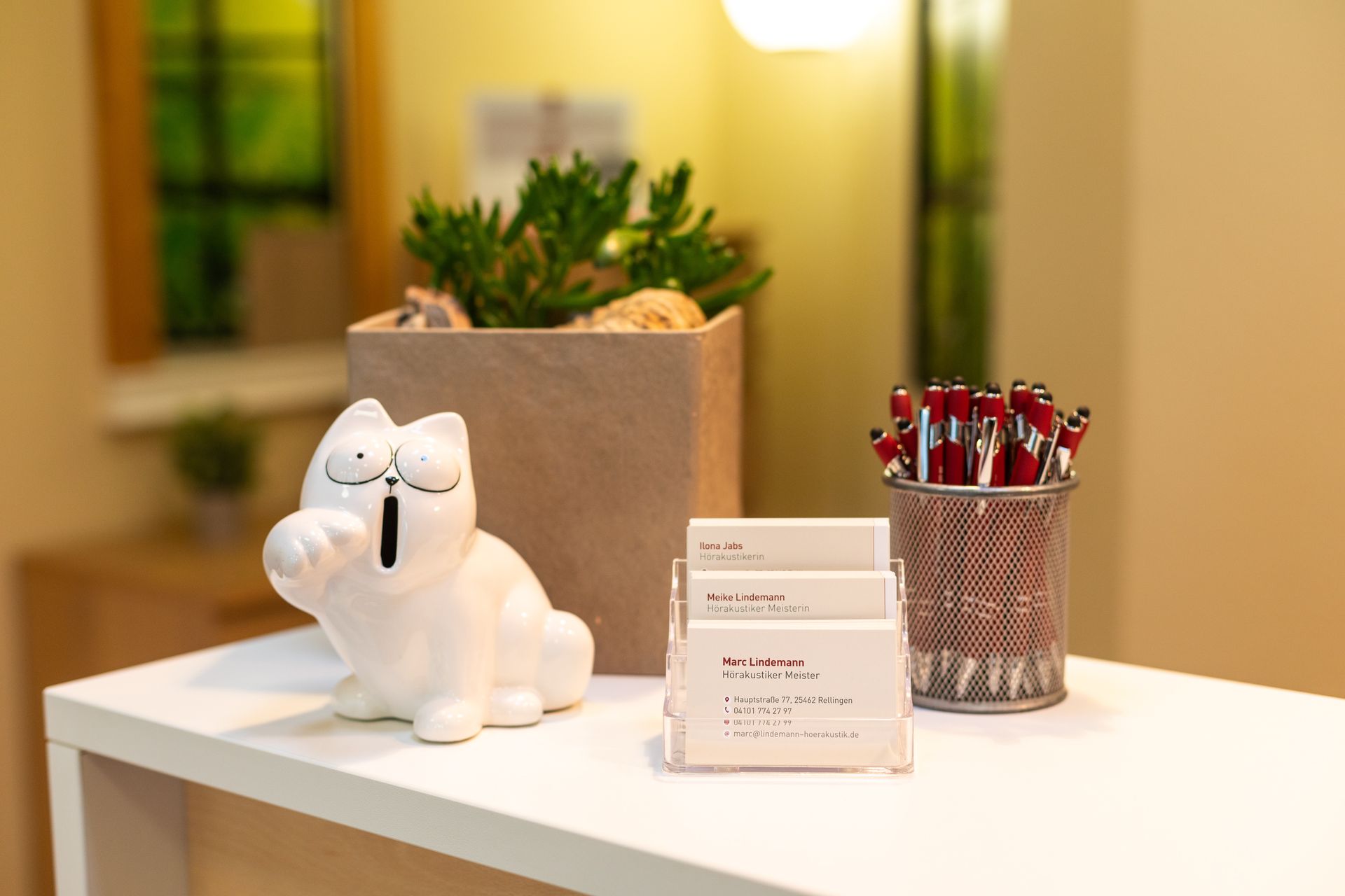 Eine weiße Katzenfigur sitzt neben einem Visitenkartenhalter auf einem weißen Tisch.
