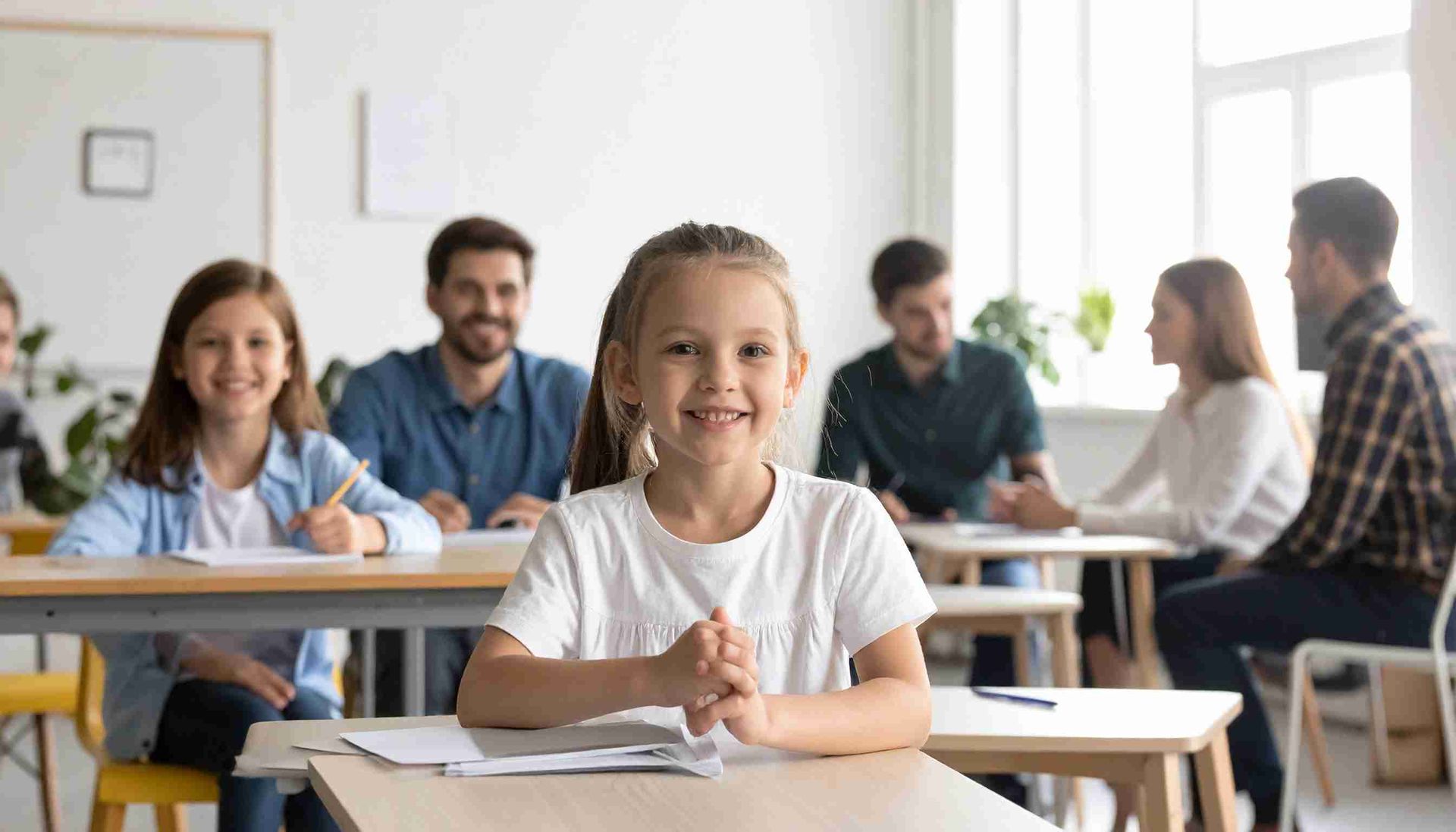 Niña sonriente en un aula con alumnos y profesores. La luz del sol se filtra por las ventanas.