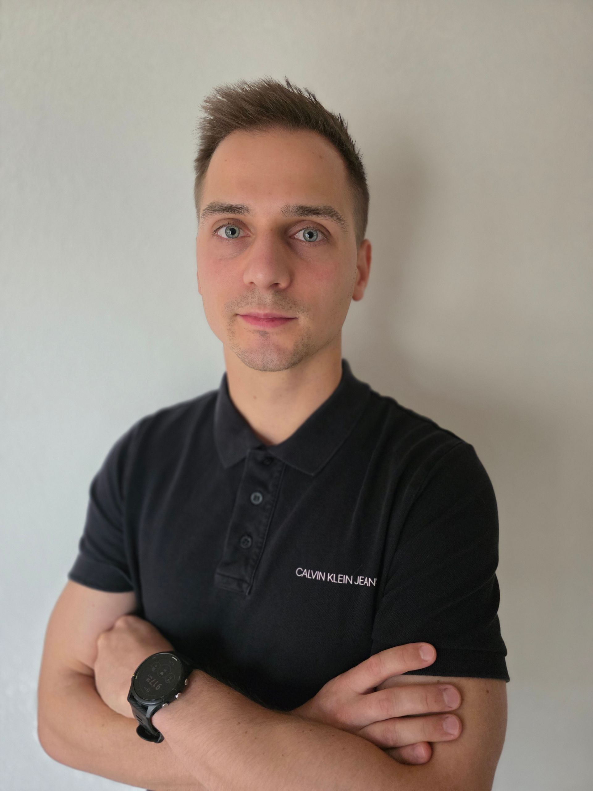 Homme aux cheveux bruns courts, portant un polo noir, les bras croisés, regardant l'objectif.