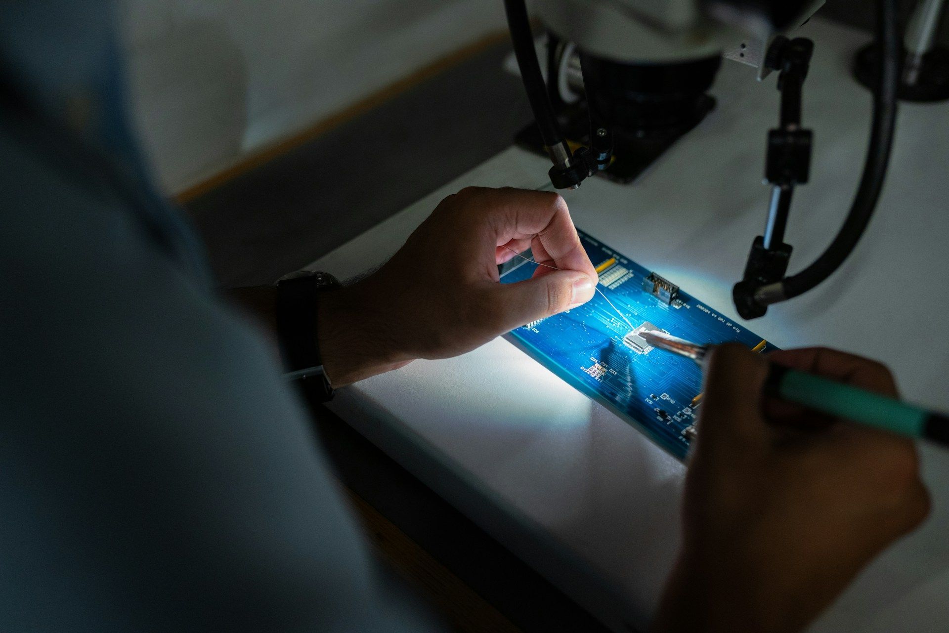 Des mains soudent un circuit imprimé bleu sous la lumière d'un microscope.