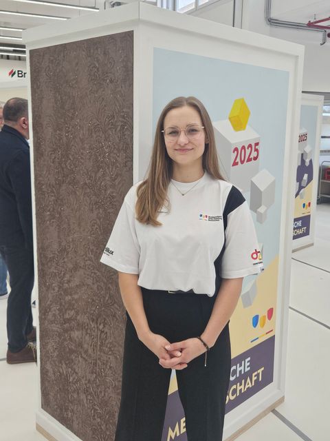 Eine Frau steht an einem Messestand, sie trägt ein weißes T-Shirt und eine schwarze Hose; hinter ihr ist ein Schild mit der Jahreszahl 2025 zu sehen.