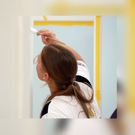 Eine Person streicht mit einem kleinen Pinsel eine weiße Wand; die Aufnahme stammt von hinten und zeigt einen hellen Raum.