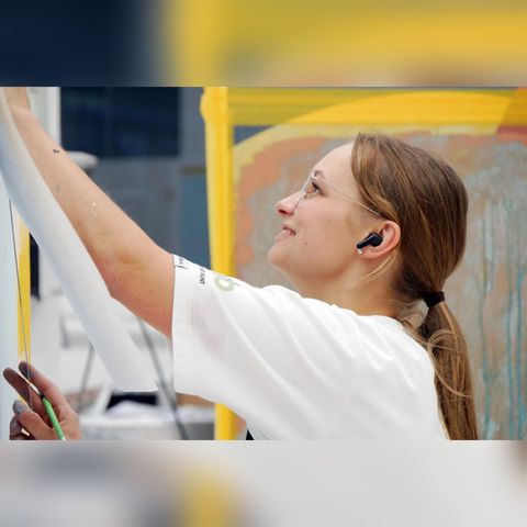 Eine Frau in weißem Hemd benutzt ein Smartphone in der Nähe eines gelben Gebäudes, lächelt und streckt die Hand nach oben.