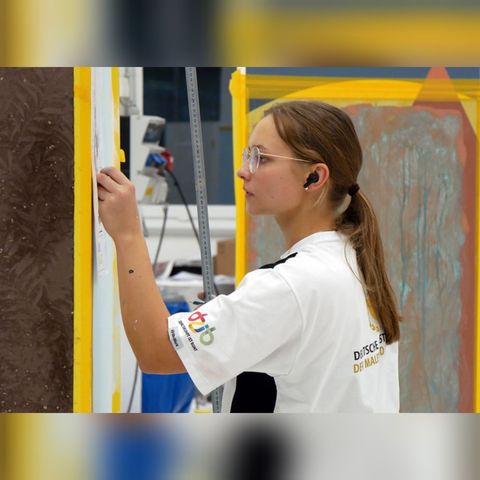 Eine Person bemalt in einer Werkstatt eine gelb umrandete Tafel; sie trägt eine Schutzbrille und Ohrstöpsel.