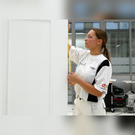 Eine Frau in weißer Arbeitskleidung streicht eine weiße Wand in einem hellen Raum.