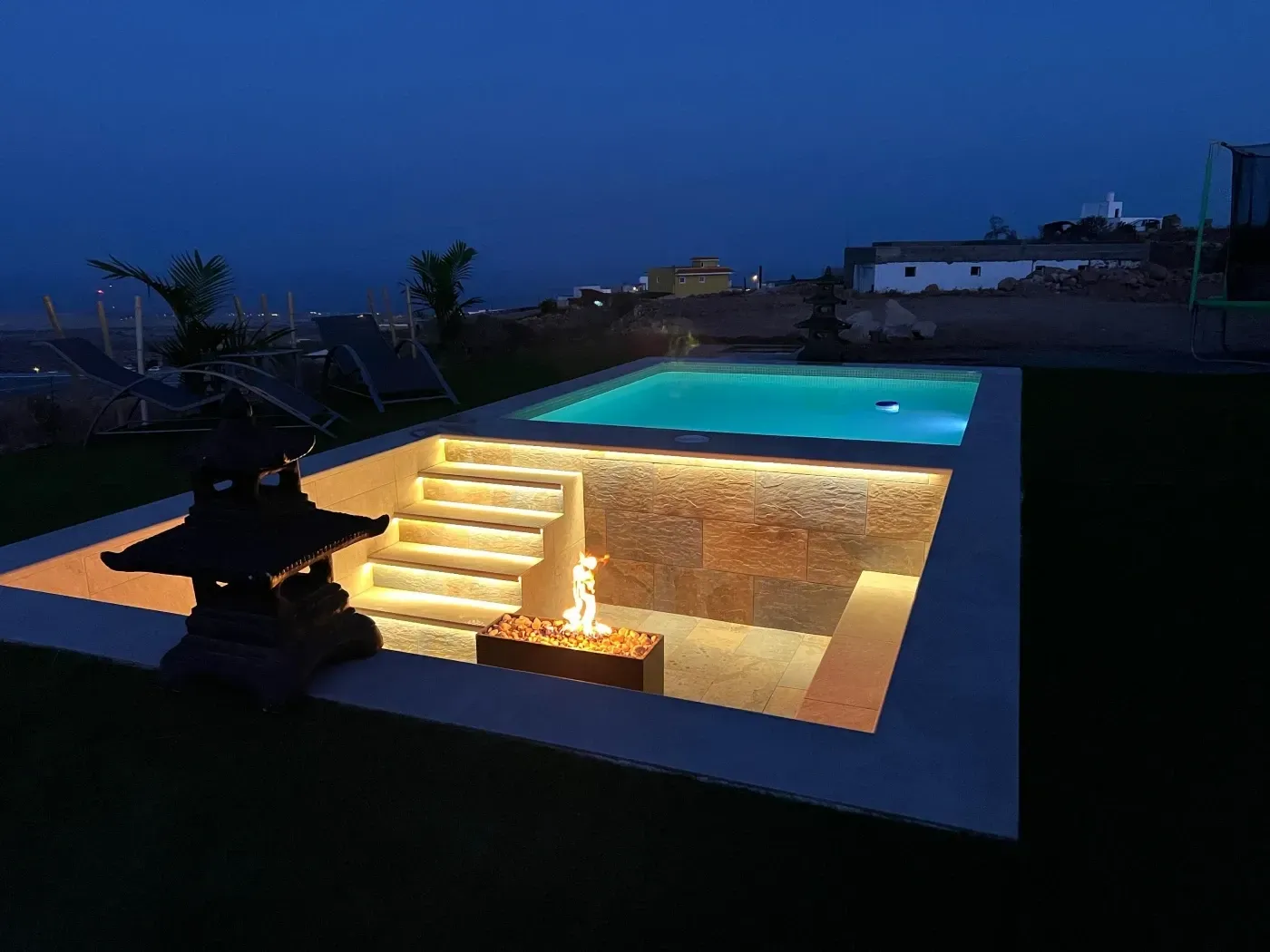 Escena nocturna: Piscina iluminada con fogata hundida y escalones de piedra