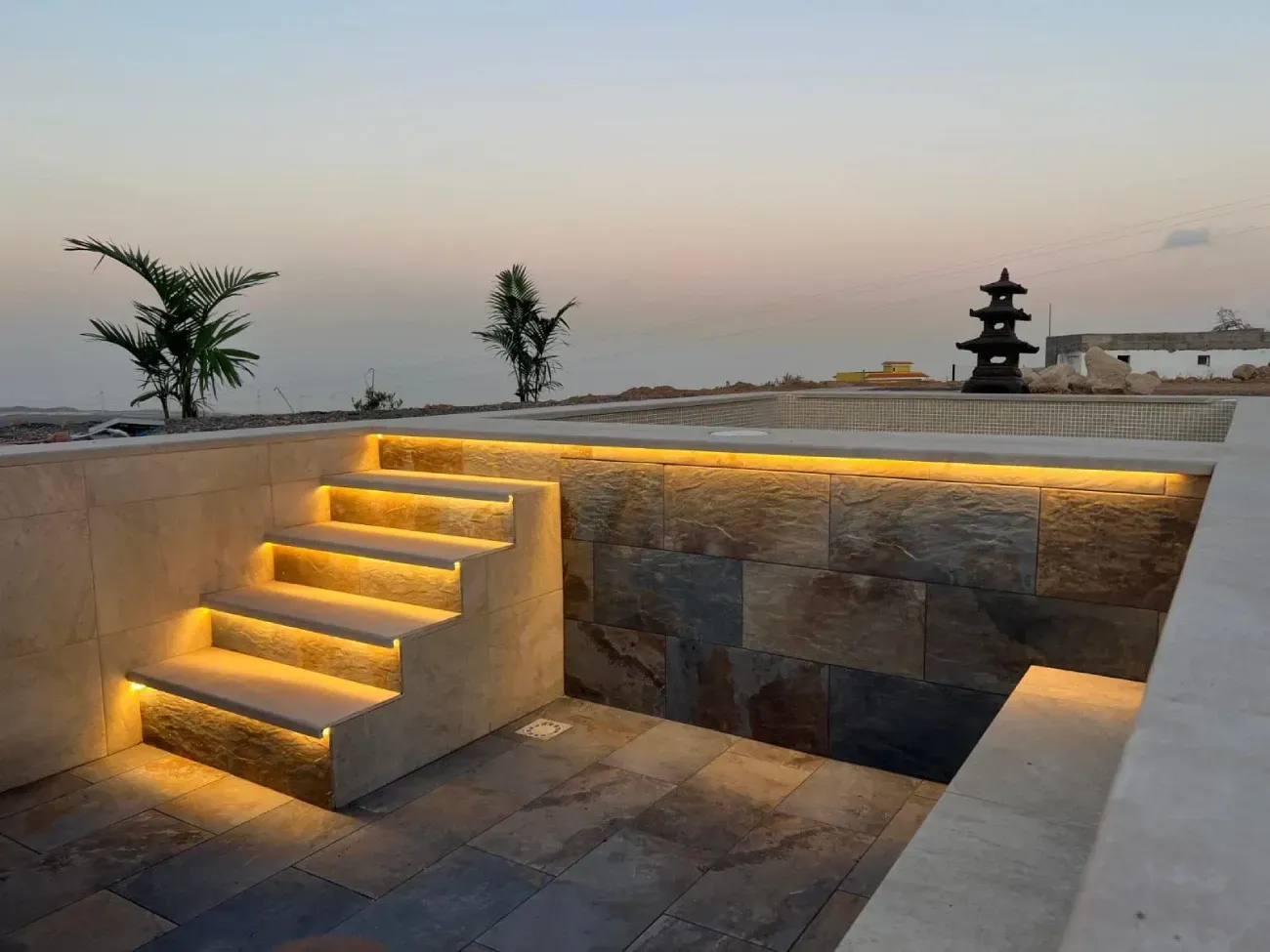 Área hundida al aire libre con escalones iluminados, muro de piedra y escultura de pagoda al atardecer.