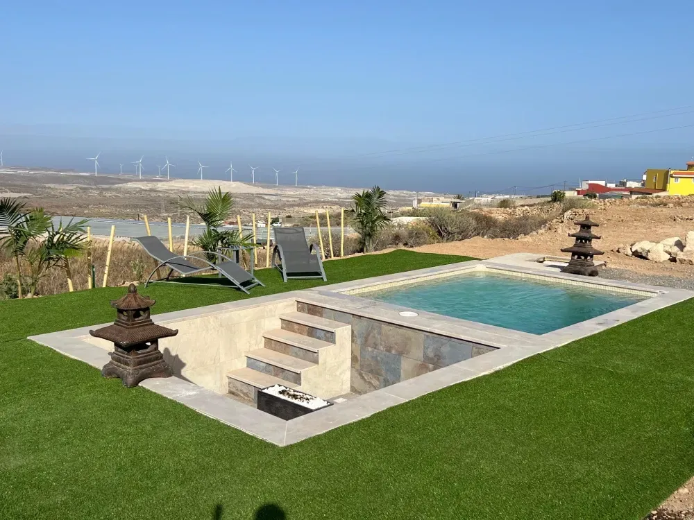 Piscina con escalones, rodeada de césped artificial y faroles de piedra. 