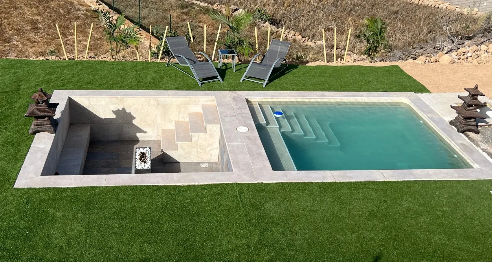 Una piscina rectangular con jacuzzi incorporado y escalones. Dos tumbonas descansan sobre el césped verde circundante.