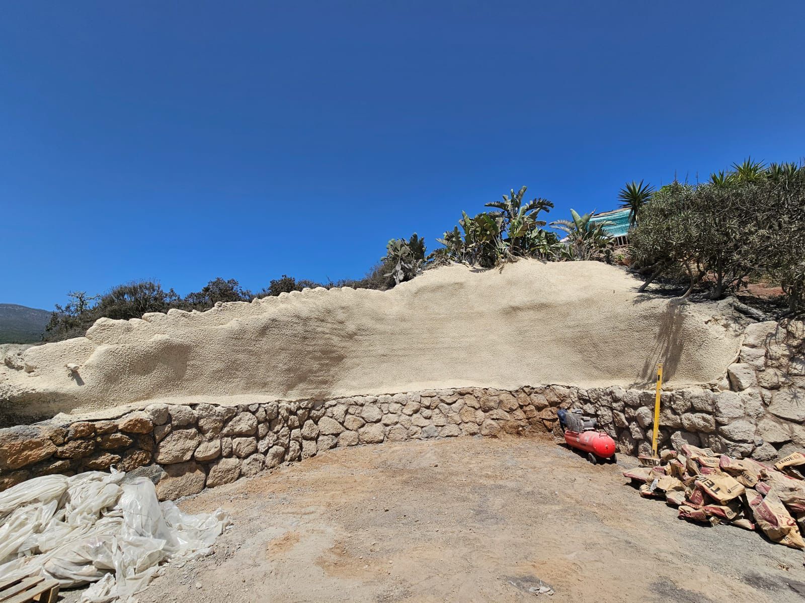 Obra con un muro de contención de piedra apilada. Arriba, bajo un cielo azul, se alza una gran pila de material claro.