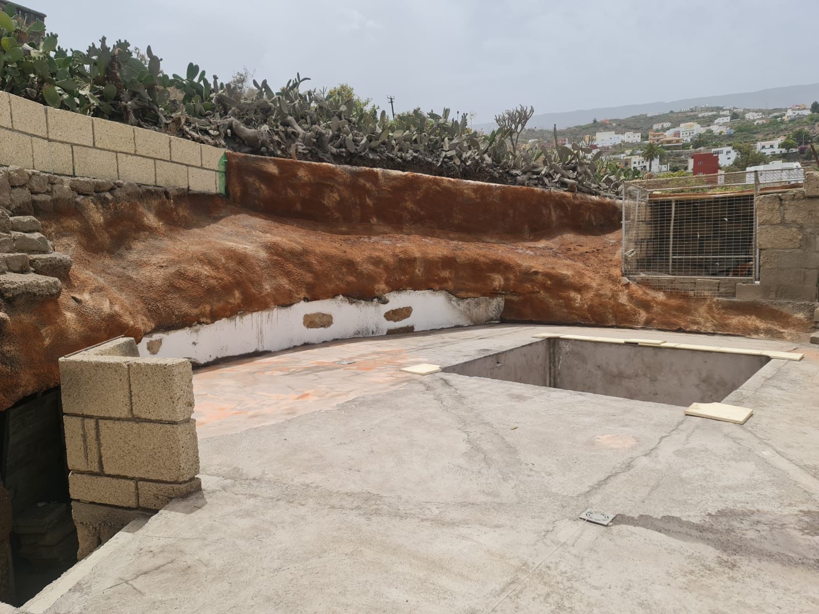 Patio de hormigón en construcción junto a un terraplén elevado de tierra con chumberas. 